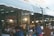 A busy market scene with several vendors and shoppers. Stalls are filled with various goods and products, and there are bright lights creating a warm, lively atmosphere. Signs with text in a Southeast Asian script hang from the stalls, adding to the market ambiance.