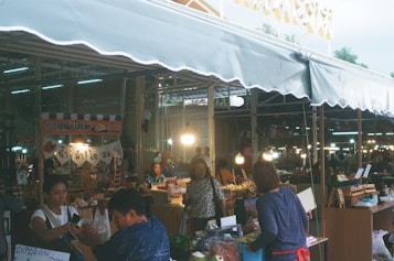 A busy market scene with several vendors and shoppers. Stalls are filled with various goods and products, and there are bright lights creating a warm, lively atmosphere. Signs with text in a Southeast Asian script hang from the stalls, adding to the market ambiance.