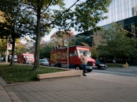 Two red food trucks are parked along a street lined with trees. A person wearing a hooded jacket and hat is walking past the trucks. The setting is urban, with a modern glass building in the background and a sidewalk in the foreground.