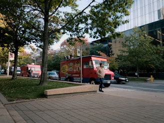 Two red food trucks are parked along a street lined with trees. A person wearing a hooded jacket and hat is walking past the trucks. The setting is urban, with a modern glass building in the background and a sidewalk in the foreground.