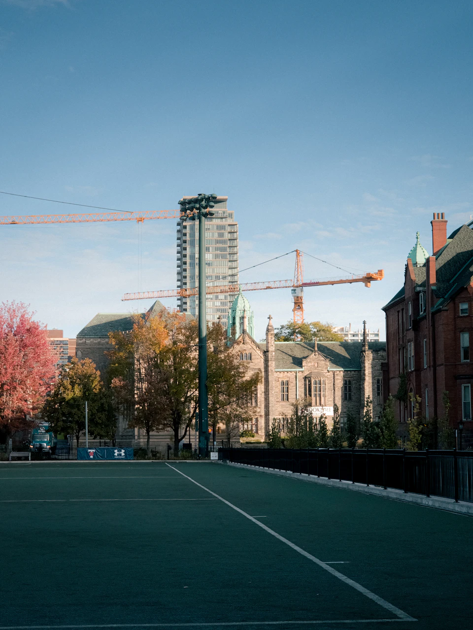 The image depicts a cityscape with a large, modern skyscraper on the right side and an older building on the left side. The sky is clear blue, and there are trees visible in the background. In the foreground, there is a tennis court with a green surface and white lines marking the boundaries of the game.