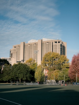 A large concrete building with modern architecture stands prominently in the background, surrounded by lush green trees with autumn foliage. In the foreground, there is an empty sports field equipped with a soccer goal and a few orange cones. The scene is bathed in daylight under a blue sky dotted with clouds.