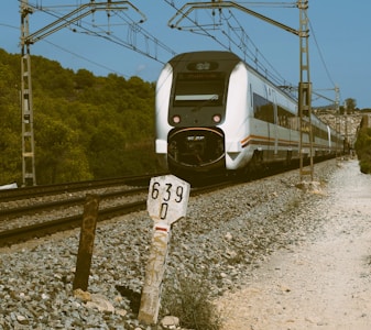 A white and black train is traveling on a pair of railway tracks with surrounding gravel and a backdrop of green trees. Overhead wires and electric poles are present, and a milestone marker labeled '639 0' stands alongside the tracks.