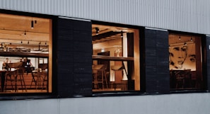 A warm and inviting interior of a modern restaurant is visible through three large windows. The space features wooden furniture, including chairs and tables, under soft ceiling lights. Artistic wall designs featuring abstract faces add a creative touch. A few people are present, possibly staff or patrons.