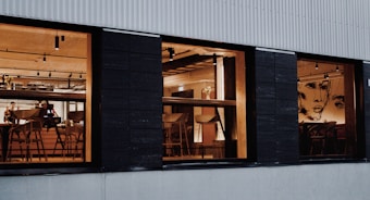 A warm and inviting interior of a modern restaurant is visible through three large windows. The space features wooden furniture, including chairs and tables, under soft ceiling lights. Artistic wall designs featuring abstract faces add a creative touch. A few people are present, possibly staff or patrons.