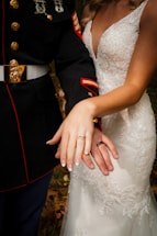 A bride and groom display their wedding rings. The groom is wearing a military uniform with medals, and the bride is wearing a lace wedding dress.
