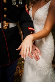 A bride and groom display their wedding rings. The groom is wearing a military uniform with medals, and the bride is wearing a lace wedding dress.