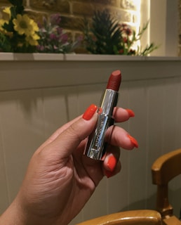 A person holds a silver-cased red lipstick in their hand, with matching nail polish. The background features a white wooden panel and a beige brick wall adorned with various flowers and greenery.