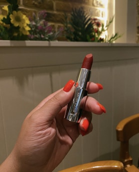 A person holds a silver-cased red lipstick in their hand, with matching nail polish. The background features a white wooden panel and a beige brick wall adorned with various flowers and greenery.