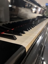 A close-up view of a piano keyboard, highlighting the black and white keys. The focus is on the glossy texture of the piano surface, with a blurred background adding depth to the image.