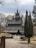 An ornate wooden church with intricate carvings and multiple steep gabled roofs, surrounded by bare trees in a natural setting. A tall, narrow tree and a stump flank the gravel path leading to the building.
