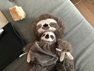 A plush toy of a sloth is laying on a grey fabric surface. The toy appears to be holding a smaller sloth toy on its chest. The background includes a piece of bread and some black fabric.