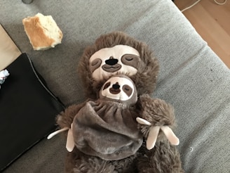 A plush toy of a sloth is laying on a grey fabric surface. The toy appears to be holding a smaller sloth toy on its chest. The background includes a piece of bread and some black fabric.