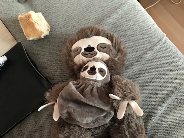 A plush toy of a sloth is laying on a grey fabric surface. The toy appears to be holding a smaller sloth toy on its chest. The background includes a piece of bread and some black fabric.
