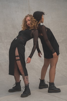 Two individuals are posing creatively against a concrete backdrop. Both are wearing black outfits featuring a blend of mesh and solid fabric pieces. Their lower halves are completed with fishnet stockings and black boots. The scene exudes a sense of style and expression.