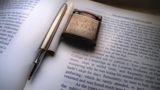 A detailed view of a printed page from a book or magazine with a lighter and a silver pen placed on top. The lighter has ornate engravings on its surface, while the pen is sleek and metallic. The text visible is in English and seems to discuss scientific or philosophical topics.