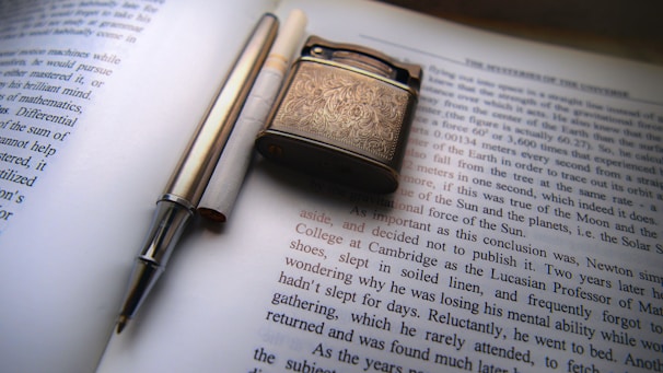 A detailed view of a printed page from a book or magazine with a lighter and a silver pen placed on top. The lighter has ornate engravings on its surface, while the pen is sleek and metallic. The text visible is in English and seems to discuss scientific or philosophical topics.