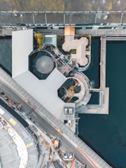 An aerial view of an architectural complex next to the water, featuring several connected modern buildings and a circular structure. The design incorporates geometric shapes and includes a visible pathway and some construction equipment nearby.