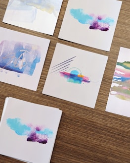 Five small pieces of paper featuring abstract watercolor designs are arranged on a wooden surface. The designs include a mix of soft pastel colors, with some featuring bold lines or artistic smudges.