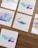Five small pieces of paper featuring abstract watercolor designs are arranged on a wooden surface. The designs include a mix of soft pastel colors, with some featuring bold lines or artistic smudges.