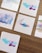 Five small pieces of paper featuring abstract watercolor designs are arranged on a wooden surface. The designs include a mix of soft pastel colors, with some featuring bold lines or artistic smudges.