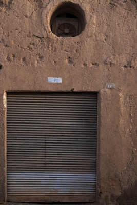 A textured, worn wall made of rough material with a large, closed metallic rolling shutter. Above the shutter, a circular opening houses an exhaust fan. Small patches of light and shadow play across the surface, indicating the time of day.