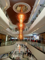 A large, elaborate chandelier with a fiery design is suspended from the ceiling of a modern, multi-level shopping mall. The chandelier's orange and yellow hues stand out amidst the clean, white lines of the mall's architecture. Crowds of people can be seen walking along the balconies and corridors of different floors.