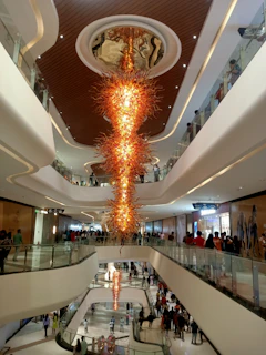 A large, elaborate chandelier with a fiery design is suspended from the ceiling of a modern, multi-level shopping mall. The chandelier's orange and yellow hues stand out amidst the clean, white lines of the mall's architecture. Crowds of people can be seen walking along the balconies and corridors of different floors.
