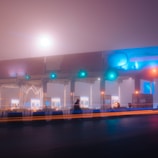 A foggy toll booth is illuminated by colorful lights in a misty atmosphere. A lone figure, silhouetted against the hazy background, stands or walks near the barriers. Light trails from passing vehicles add dynamic lines across the scene, enhancing the quiet and surreal ambiance of the setting.
