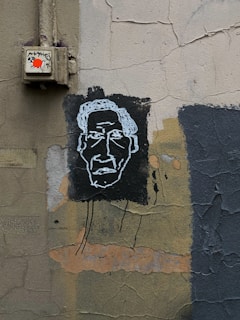 A textured wall featuring street art with a simplistic white outline of a face on a black background. The wall has an aged appearance, with cracked and peeling paint in shades of gray, green, and brown. An electrical box with a bright red sticker is mounted on the wall above the artwork.