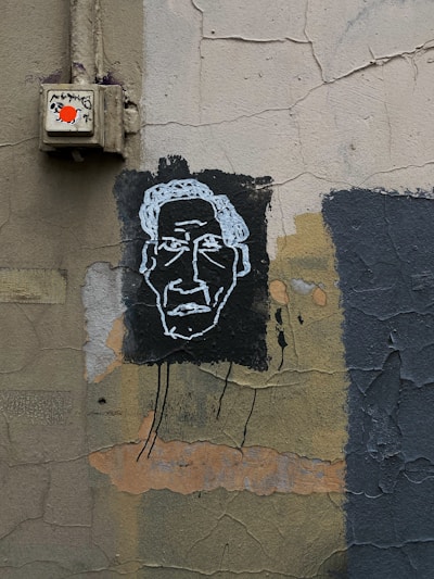 A textured wall featuring street art with a simplistic white outline of a face on a black background. The wall has an aged appearance, with cracked and peeling paint in shades of gray, green, and brown. An electrical box with a bright red sticker is mounted on the wall above the artwork.