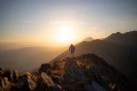 Figura solitaria en cresta de montaña caminando hacia el horizonte con luz dorada de atardecer y cadenas montañosas al fondo