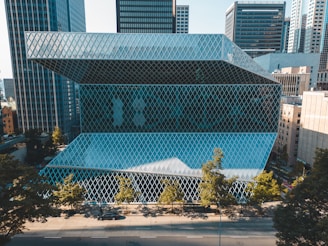 A modern glass building with a unique architectural design, featuring diamond-shaped patterns on its facade, surrounded by high-rise buildings in a cityscape. Trees line the street in front of the building, adding a touch of nature to the urban setting.