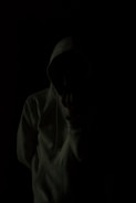 A person wearing a hoodie is standing against a very dark background, creating a mysterious and shadowy appearance. The person's face is obscured, adding to the anonymity.