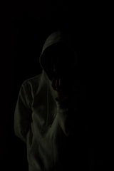 A person wearing a hoodie is standing against a very dark background, creating a mysterious and shadowy appearance. The person's face is obscured, adding to the anonymity.