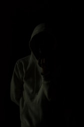 A person wearing a hoodie is standing against a very dark background, creating a mysterious and shadowy appearance. The person's face is obscured, adding to the anonymity.