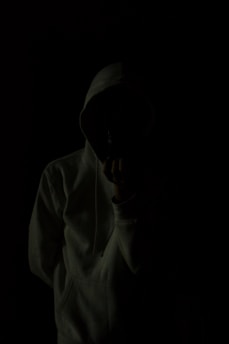 A person wearing a hoodie is standing against a very dark background, creating a mysterious and shadowy appearance. The person's face is obscured, adding to the anonymity.