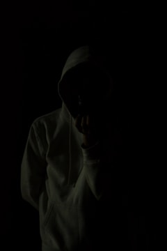 A person wearing a hoodie is standing against a very dark background, creating a mysterious and shadowy appearance. The person's face is obscured, adding to the anonymity.