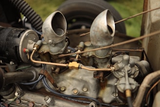A vintage car engine with visible components including two carburetors, various metal pipes, and bolts. The metal surfaces show signs of wear and oxidation, giving it a rustic appearance. The engine is surrounded by several connecting wires and hoses, hinting at its mechanical complexity.