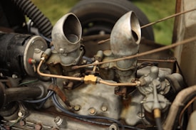 A vintage car engine with visible components including two carburetors, various metal pipes, and bolts. The metal surfaces show signs of wear and oxidation, giving it a rustic appearance. The engine is surrounded by several connecting wires and hoses, hinting at its mechanical complexity.
