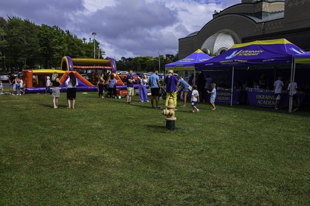 A group of people, including children, are gathered on a grassy field near inflatable play structures and tents labeled 'Ukrainian Academy'. Several people are interacting around the tents and inflatable setup. The sky is partly cloudy and the setting appears to be a park or open outdoor area.