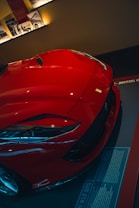 A sleek, red sports car is partially visible, showcasing its elegant design and high-performance features. The focus is on the front, highlighting its aerodynamic curves, sharp headlights, and a prominent emblem. The background includes subtle lighting and informative panels, possibly indicating a showroom or exhibit setting.