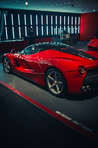 A sleek red sports car is displayed in a modern showroom with ambient lighting. The car features a stylish aerodynamic design with shiny alloy wheels. Reflective surfaces and contours emphasize its luxurious appearance. A sign on the floor says 'NON TOCCARE - DON'T TOUCH.' The backdrop includes vertical light fixtures adding to the sophisticated ambiance. A person is visible in the background, contributing to the sense of scale.
