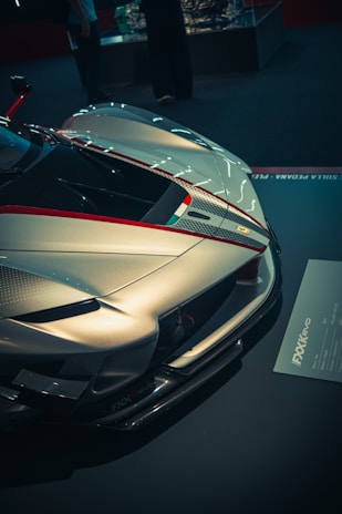 A sleek, high-performance sports car with aerodynamic features and a shiny metallic finish is showcased in an indoor setting. The car features detailed design elements, including a red stripe and high-quality carbon fiber parts. There are people in the background, possibly spectators or attendees at an event.