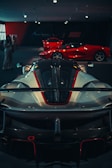 Several high-performance sports cars are displayed in a dimly lit showroom. The foreground features a sleek, aerodynamic car with a prominent spoiler and detailed bodywork. In the background, two more bright red sports cars are visible, highlighting the luxurious and exclusive nature of the collection.