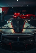 Several high-performance sports cars are displayed in a dimly lit showroom. The foreground features a sleek, aerodynamic car with a prominent spoiler and detailed bodywork. In the background, two more bright red sports cars are visible, highlighting the luxurious and exclusive nature of the collection.
