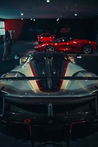 Several high-performance sports cars are displayed in a dimly lit showroom. The foreground features a sleek, aerodynamic car with a prominent spoiler and detailed bodywork. In the background, two more bright red sports cars are visible, highlighting the luxurious and exclusive nature of the collection.