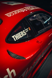 A red sports car with black details featuring a window with decals including the word 'THIERS' and a small flag. The car body displays the words 'SCUDERIA' above the window.