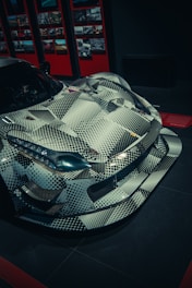 A sleek sports car is displayed, featuring a unique patterned design with black and white geometric shapes. It has an aggressive, aerodynamic shape highlighted by sharp lines and prominent features. The background includes a wall with numerous framed photographs and a predominantly dark ambiance.