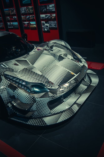 A sleek sports car is displayed, featuring a unique patterned design with black and white geometric shapes. It has an aggressive, aerodynamic shape highlighted by sharp lines and prominent features. The background includes a wall with numerous framed photographs and a predominantly dark ambiance.